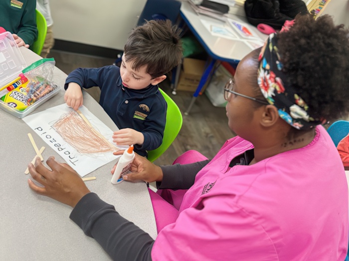 A teacher guides a child in creating art with crayons and glue, focusing on a colorful drawing in a bright classroom.