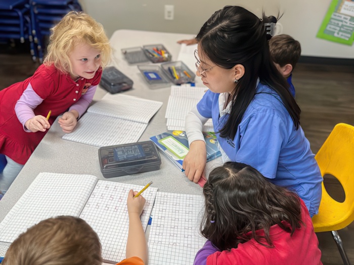 A teacher helps children with their work at a table, encouraging collaboration in a bright and engaging classroom.