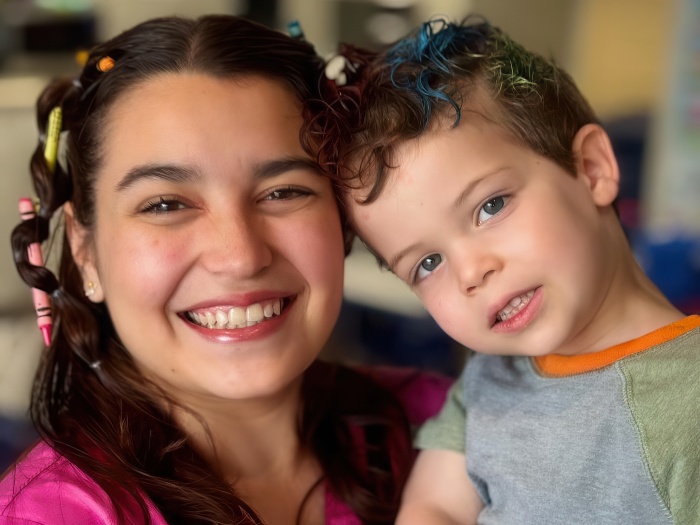 A smiling teacher with colorful hair ties poses with a cheerful young boy in a casual setting, radiating warmth and joy.