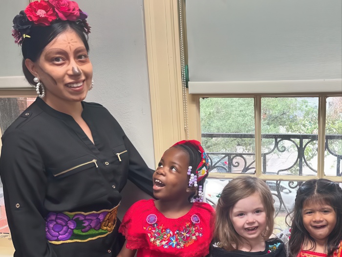 A woman in traditional attire smiles with three girls in colorful outfits, all joyful, near a window with a bright view.