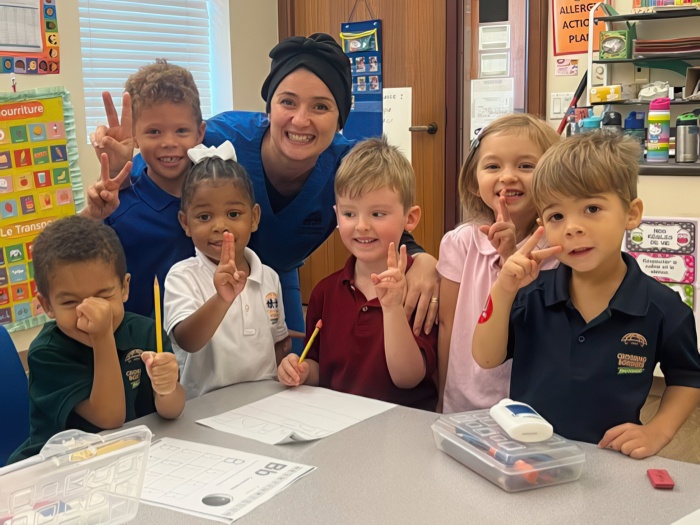 A smiling teacher poses with six children, all making peace signs and enjoying art activities in a colorful classroom.