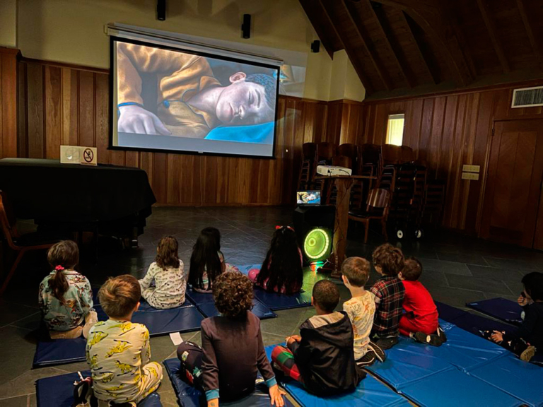 Children sit on mats, attentively watching a movie projected on a large screen in a cozy indoor space.