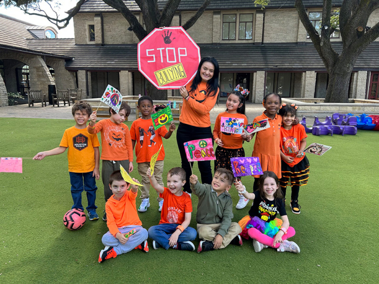 A group of children in orange shirts and their teacher hold anti-bullying signs in a colorful outdoor setting.