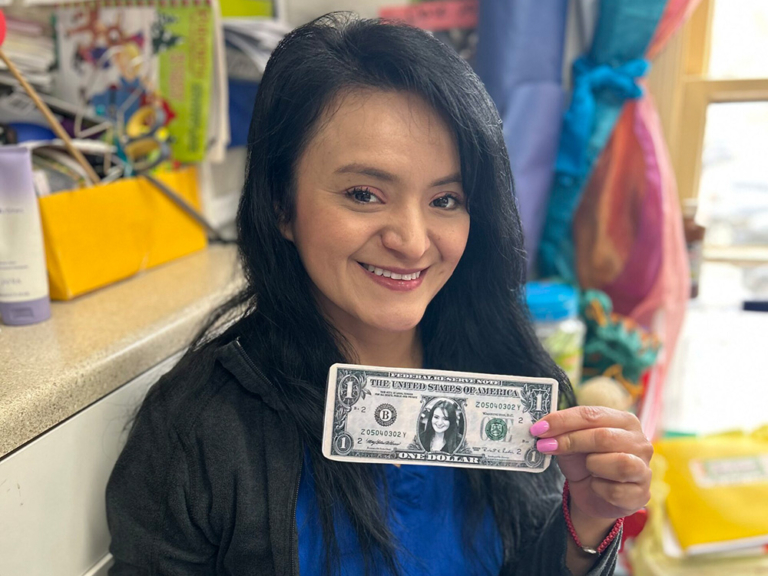 A woman smiles while holding a one-dollar bill, sitting at a desk surrounded by colorful items.