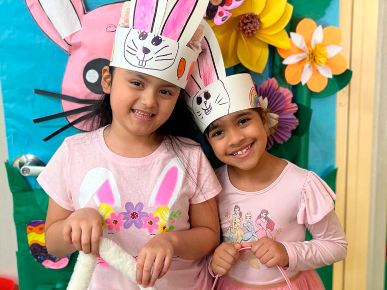 Two girls wearing bunny hats smile together, holding fluffy tails, in front of a colorful spring-themed backdrop.