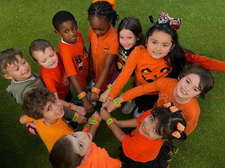 A group of children in orange shirts form a circle, smiling and stacking their hands together on a green surface.