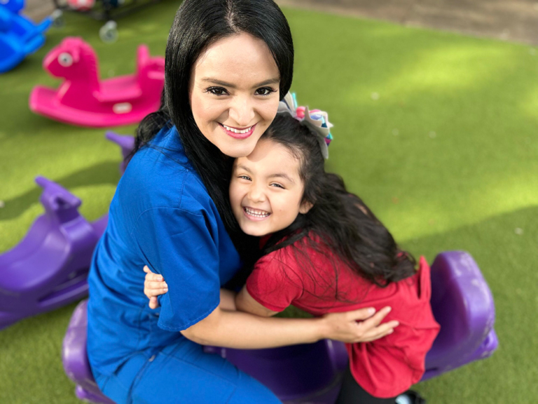 A teacher and a girl smile joyfully while sitting on a purple ride-on toy, enjoying a playful moment together outdoors.