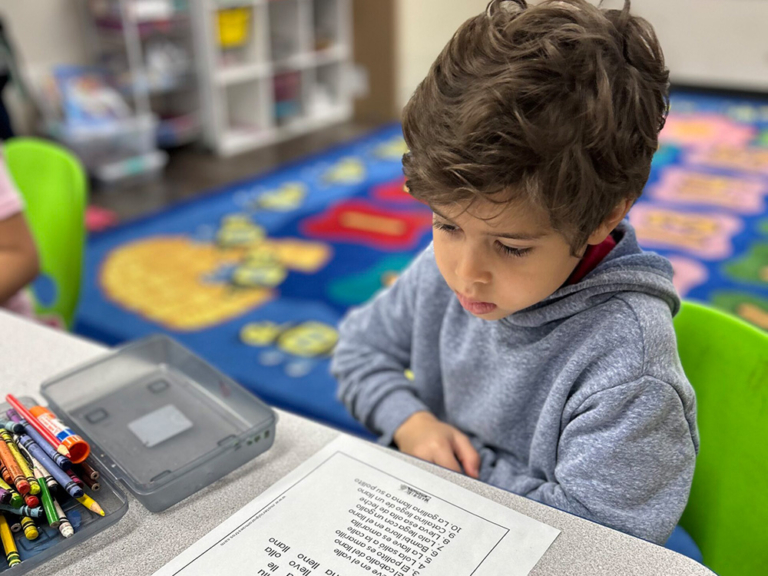 A young boy in a gray hoodie focuses intently on a worksheet at a table, surrounded by colorful crayons and a vibrant carpet.