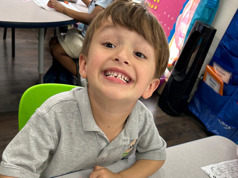 A cheerful boy in a gray shirt grins widely at the camera while sitting at a table, radiating joy and enthusiasm.