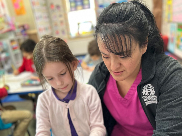 A teacher and a girl work closely together at a table, focused on an activity in a colorful classroom setting.