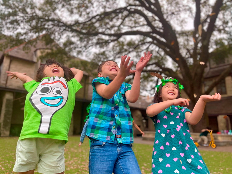 Three children playfully reach for something in the air, joyfully laughing outdoors under a large tree.