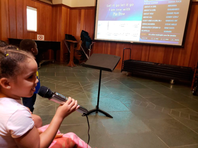 A girl holds a microphone, singing along to lyrics on a screen in a cozy indoor setting, filled with musical equipment.