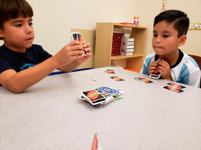 Two boys focus intently on a card game, holding up their cards at a table filled with colorful game pieces.