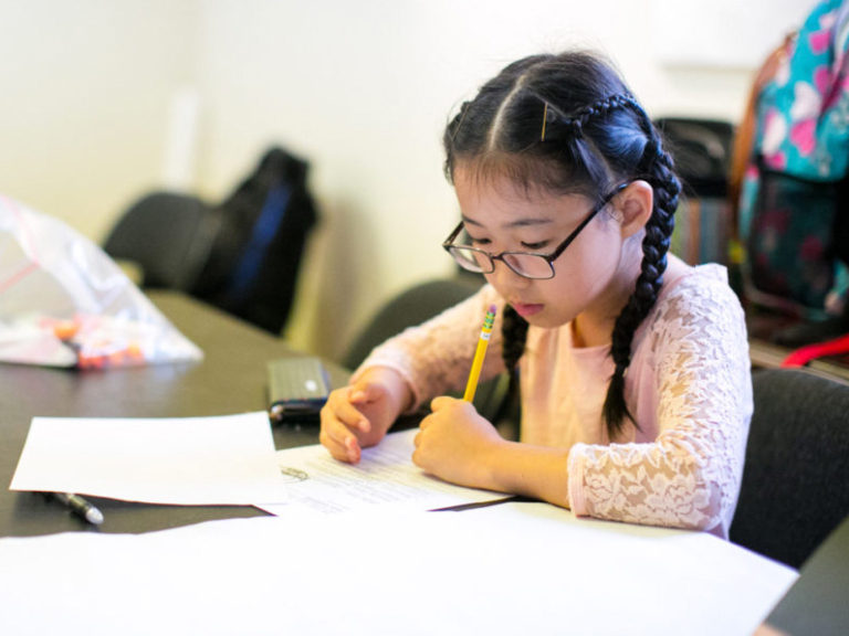 A girl with glasses focuses intently on writing, surrounded by papers and school supplies at a table.